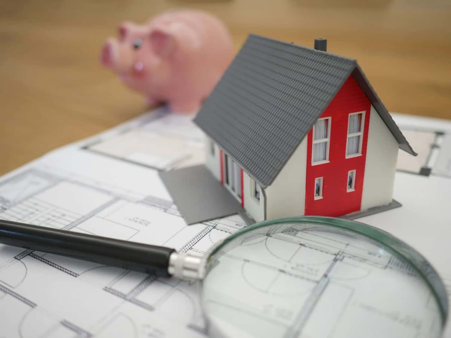 Um modelo de casa em miniatura colocado sobre uma planta de construção, com uma lupa e um cofrinho ao lado
