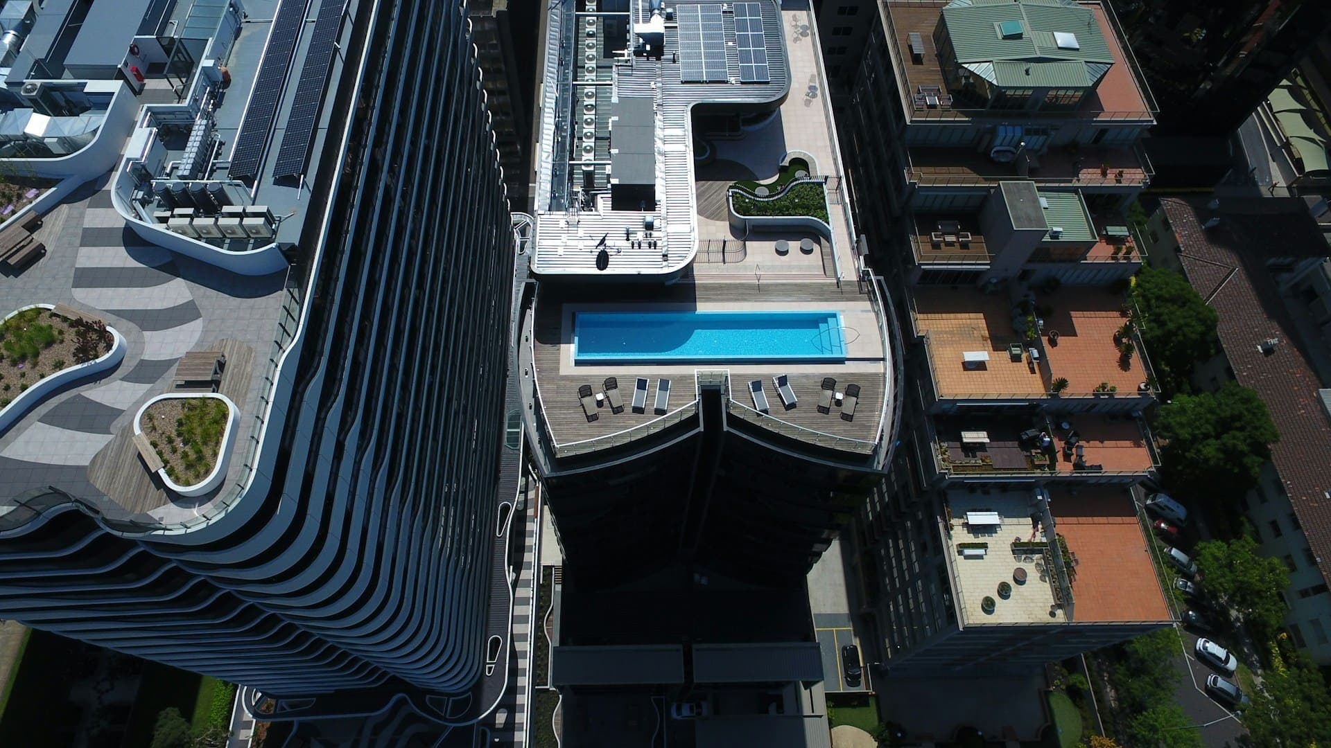 Vista aérea de uma piscina na cobertura de prédios modernos, cercada por espreguiçadeiras e áreas de convivência, em meio a uma paisagem urbana com construções residenciais e comerciais.